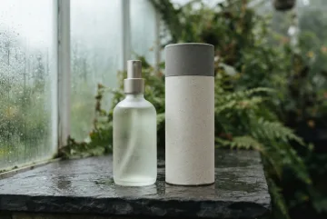 Frosted glass room spray bottle and cylindrical paper canister on a rain-darkened stone shelf in a misty conservatory.