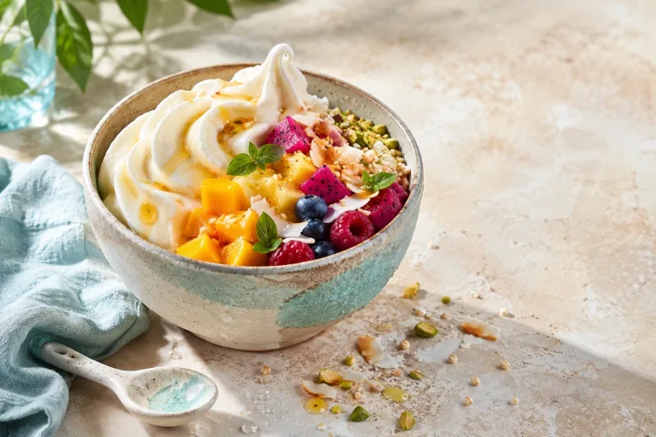 Creamy frozen yogurt topped with tropical fruit, toasted coconut, puffed grains, pistachios, basil leaves, and amber syrup on a bright stone table.