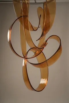 Translucent amber resin ribbons suspended in graceful abstract curves under soft studio lighting against a warm gray background.