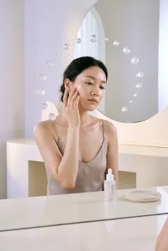Surreal beauty model applying facial serum while translucent pearl-like droplets float around her face above a reflective vanity.