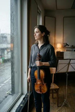 Young violinist holding a wooden violin beside a rain-streaked rehearsal room window in a cinematic portrait.