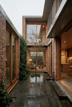 Contemporary brick courtyard house with a vertical garden, wet stone paving, concrete overhangs, timber windows, and warm interior lights during gentle rain.