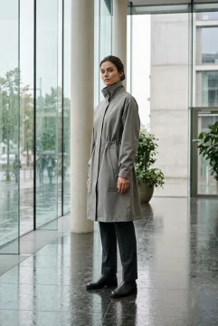 Fashion model wearing a stone-gray tailored raincoat in a misty glass atrium with rain-streaked windows and reflective stone flooring.