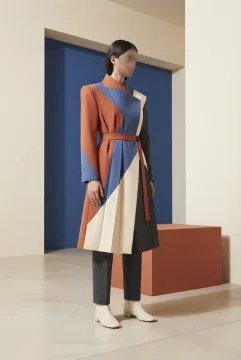 Anonymous fashion model wearing a terracotta, cobalt, ivory, and charcoal origami-pleat coat in a minimalist geometric gallery with angular shadows and a rectangular pedestal.