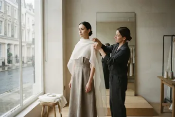 Anonymous fashion model wearing a translucent ivory organza cape over a sculptural taupe linen dress while a stylist adjusts the shoulder drape inside a minimalist atelier illuminated by soft rainy window light.