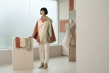 Model wanita anonim mengenakan cape geometris terracotta, ivory, dan sage dengan lipatan segitiga serta panel trapesium di atelier fashion modern bergaya Graphic Geometric.