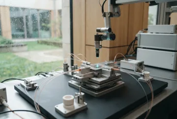 Fotografi editorial laboratorium kalibrasi chip fotonik dengan modul semikonduktor perak, serat optik transparan, dan lengan robot presisi di atas meja pengujian, berlatar jendela berkabut hujan.