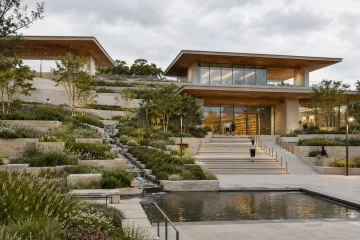 Terraced public learning pavilion with rain gardens, reflective water channels, pale stone walls, timber ceilings, and softly illuminated study rooms.