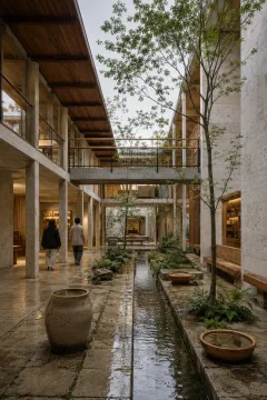 Portrait view of a contemporary civic archive pavilion with pale masonry, timber galleries, rain-darkened courtyard paving, reflective water channels, native planting, and two anonymous visitors.