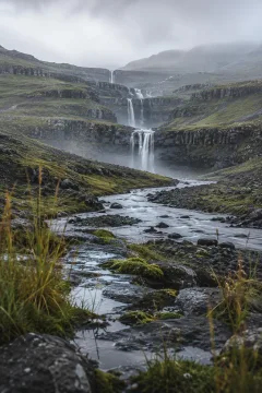 Portrait view of a misty rain-washed basalt valley with slender waterfalls flowing into a winding river between dark wet terraces and muted green highlands.