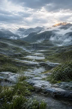 Vertical post-rain mountain valley with pale stone terraces, silver-green grasses, reflective pools, drifting mist, layered mountains, and a distant waterfall beneath luminous clouds.