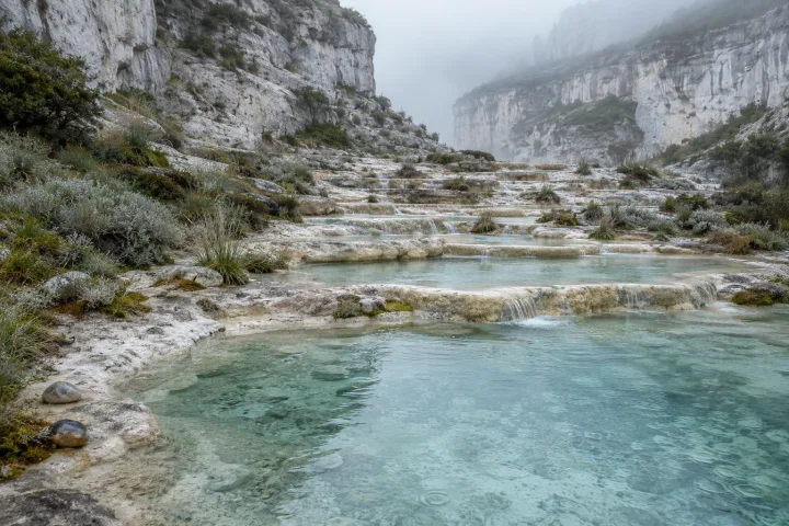 Rain-fresh pale limestone terraces descending into tranquil blue-green mineral lagoons beneath mist-softened cliffs and muted alpine vegetation.