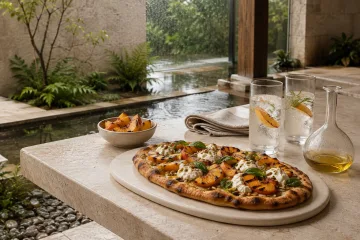Charred peach flatbread with creamy cheese and basil beside sparkling botanical drinks on a pale stone courtyard table during gentle rain.