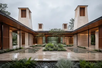 Rain-washed contemporary limestone courtyard pavilion with four sculptural windcatcher towers, timber louvers, sheltered walkways, shallow cooling basins, and restrained greenery.