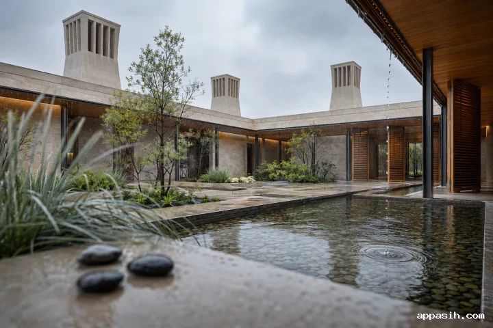 Closer three-quarter view of a rain-washed limestone windcatcher courtyard pavilion with four passive-cooling towers, timber louvers, planted walkways, and a rippling shallow water basin.