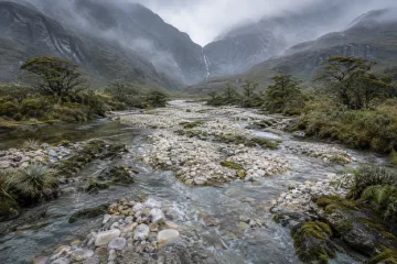 Rain-washed braided mountain river flowing among pale quartz gravel bars, mossy rock shelves, silver-green grasses, and misty blue-gray ridges.