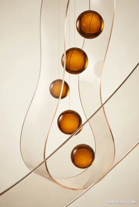 Closer abstract studio view of five amber glass spheres suspended within translucent folded acrylic ribbons under soft directional lighting.