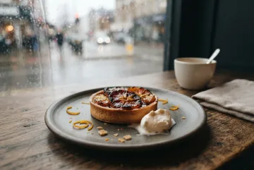 Cinematic charred citrus tart with caramelized orange slices and cream on a dark café table beside a rain-streaked window.